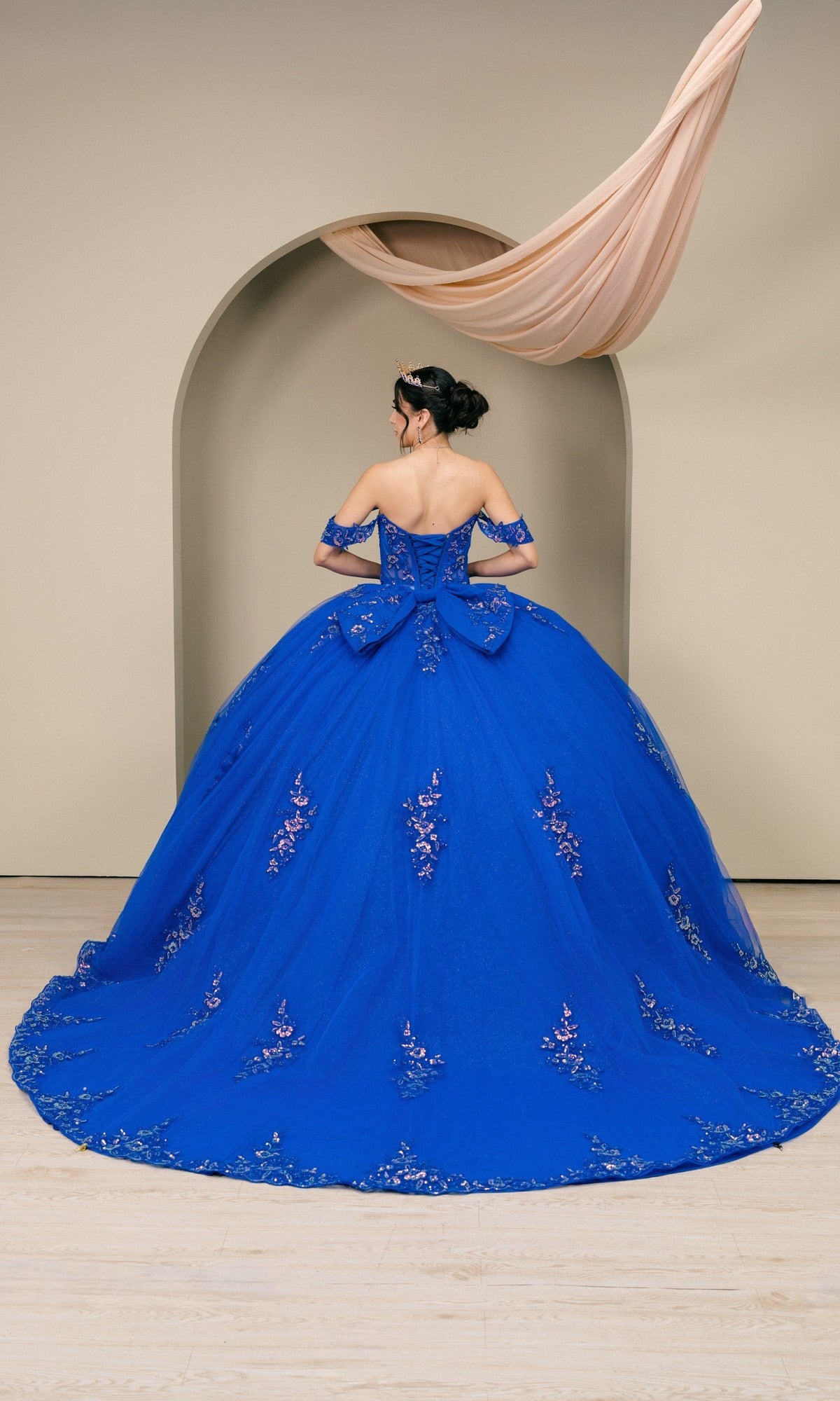 A woman stands with her back to the camera, wearing the Quinceanera Dress 8405JQ By Cinderella Couture—an off-the-shoulder royal blue ball gown with floral embroidery and a large bow—before an arched backdrop.